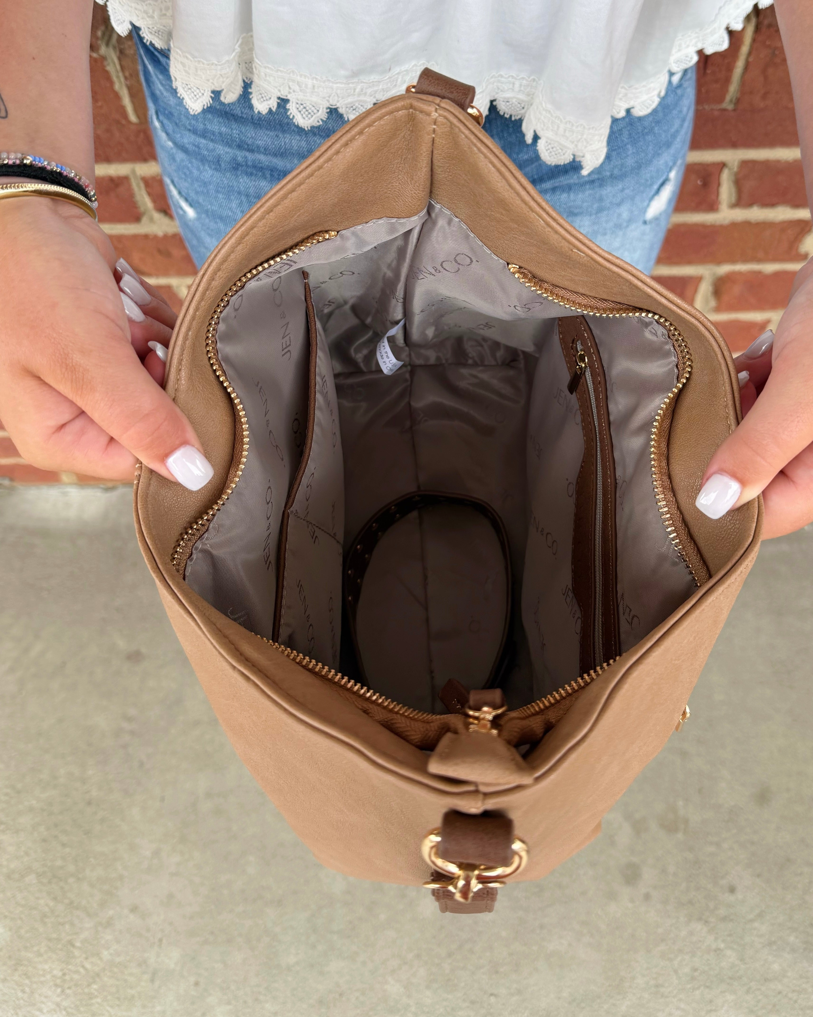 A tan bucket bag with a gold-tone clasp, held open by a person's hands to show the interior lining and zip compartments.