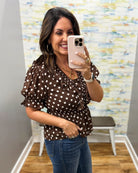 A woman takes a mirror selfie in a fitting room wearing a brown short-sleeve blouse with white polka dots and blue jeans.