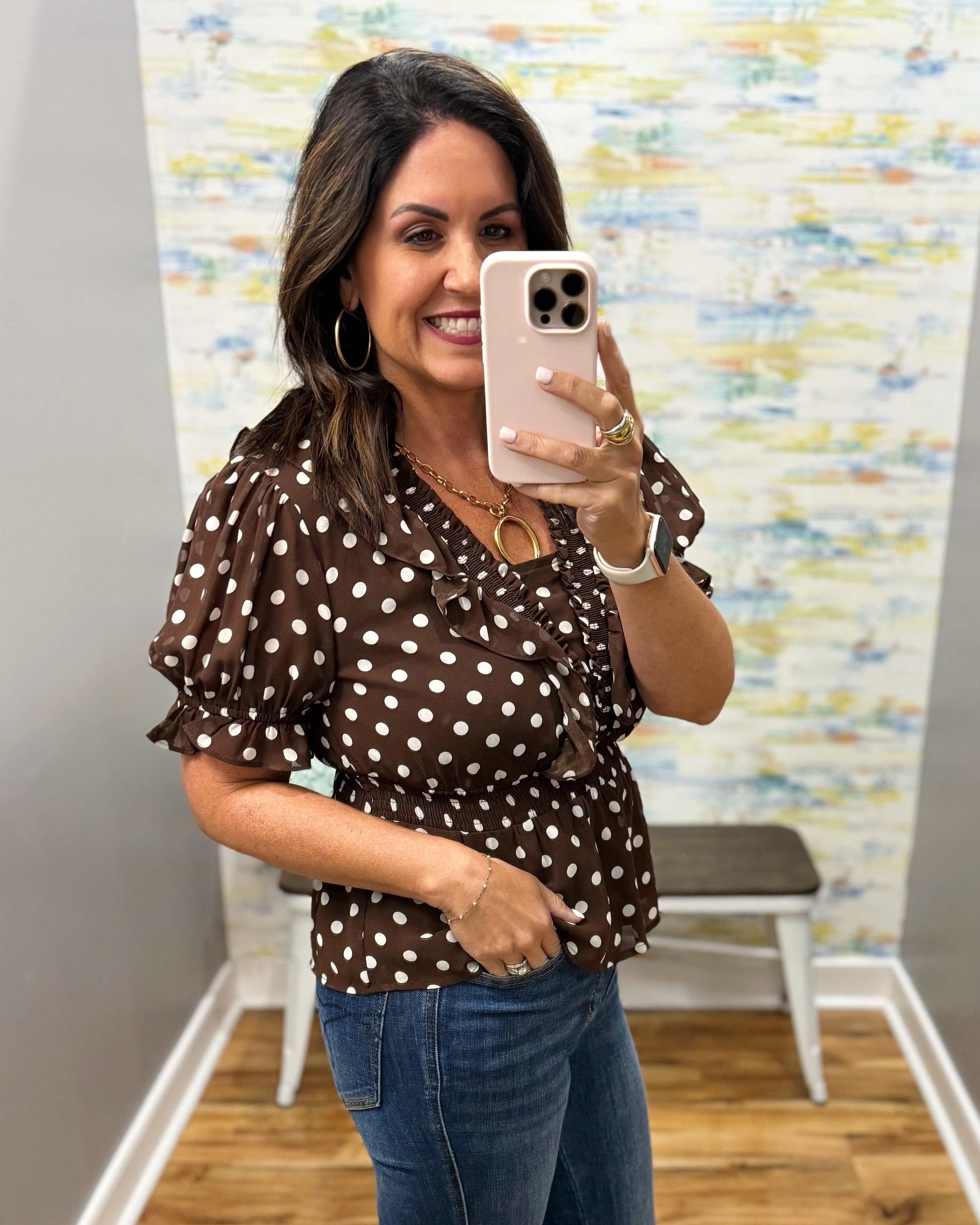 A woman takes a mirror selfie in a fitting room wearing a brown short-sleeve blouse with white polka dots and blue jeans.