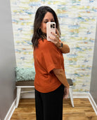 A woman takes a mirror selfie wearing a short-sleeve knit top in burnt orange and black wide-leg pants, standing in a room with a patterned wallpaper background.