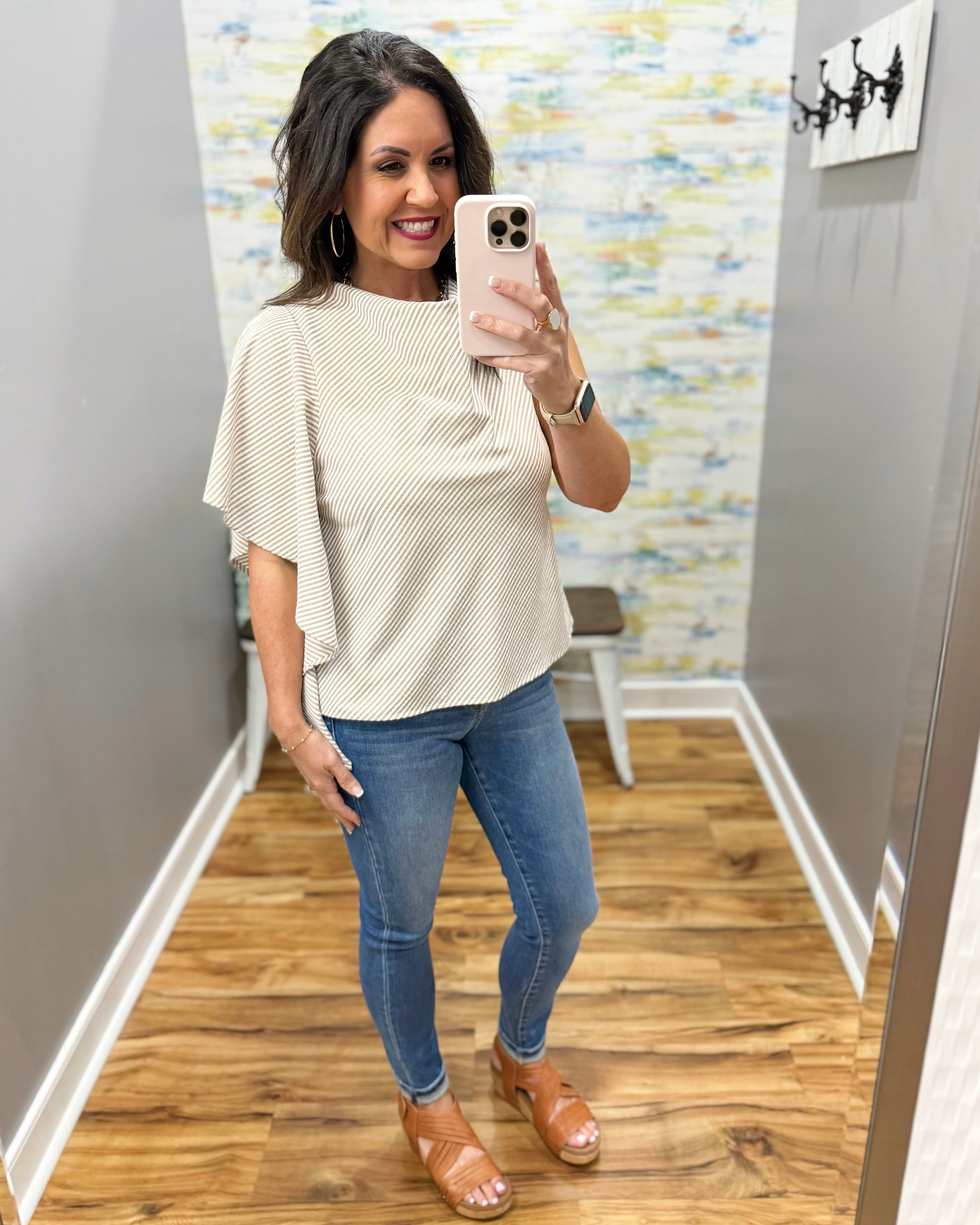 Woman taking a mirror selfie wearing a beige top and blue jeans in a room with a patterned wall.