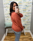 A woman takes a mirror selfie wearing a rust-orange long-sleeve knit top and light-wash blue jeans in a room with a patterned wallpaper background.