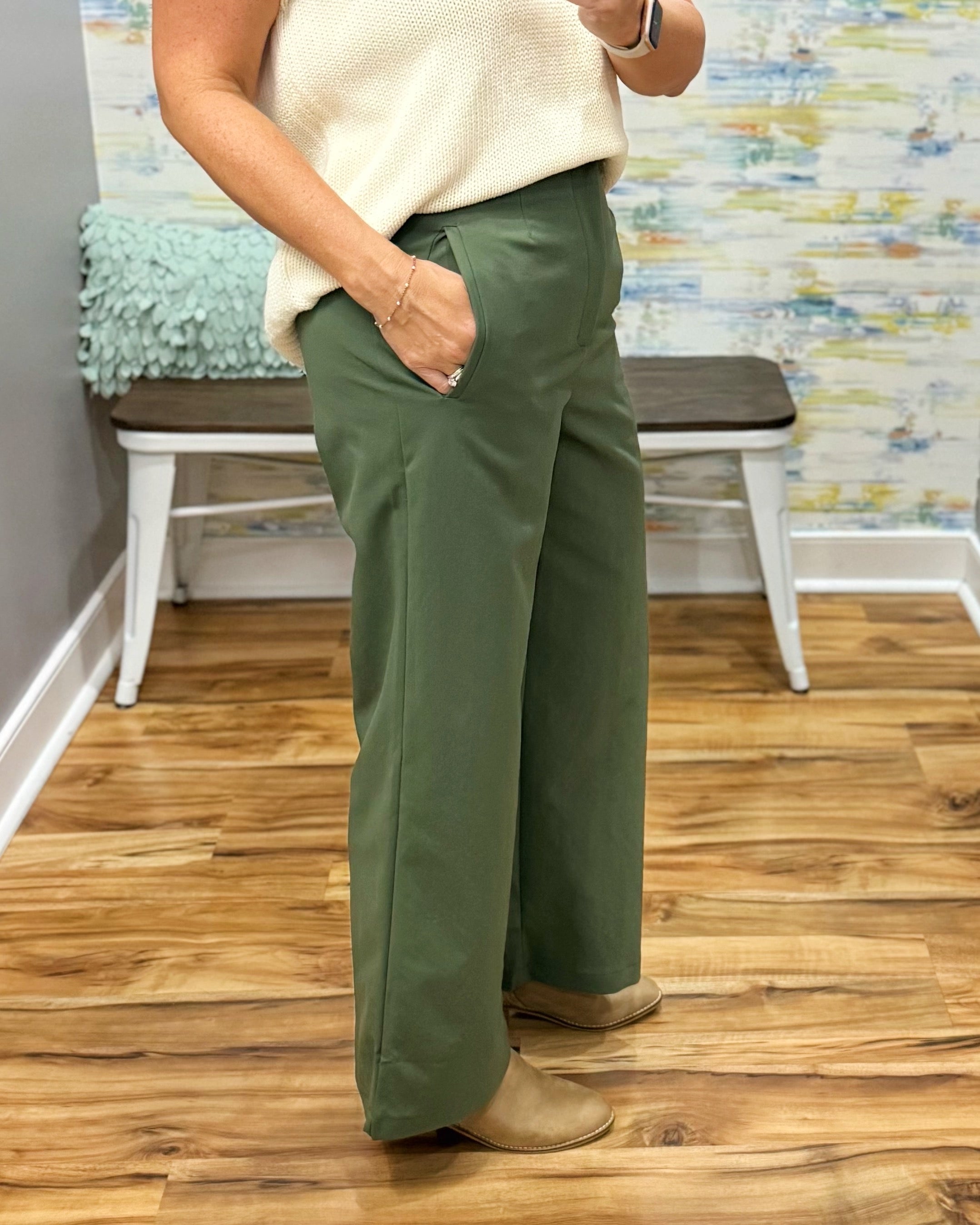 A person wearing olive green wide-leg pants, a cream knit top, and tan shoes, standing in a room with a wood floor and a bench in the background.