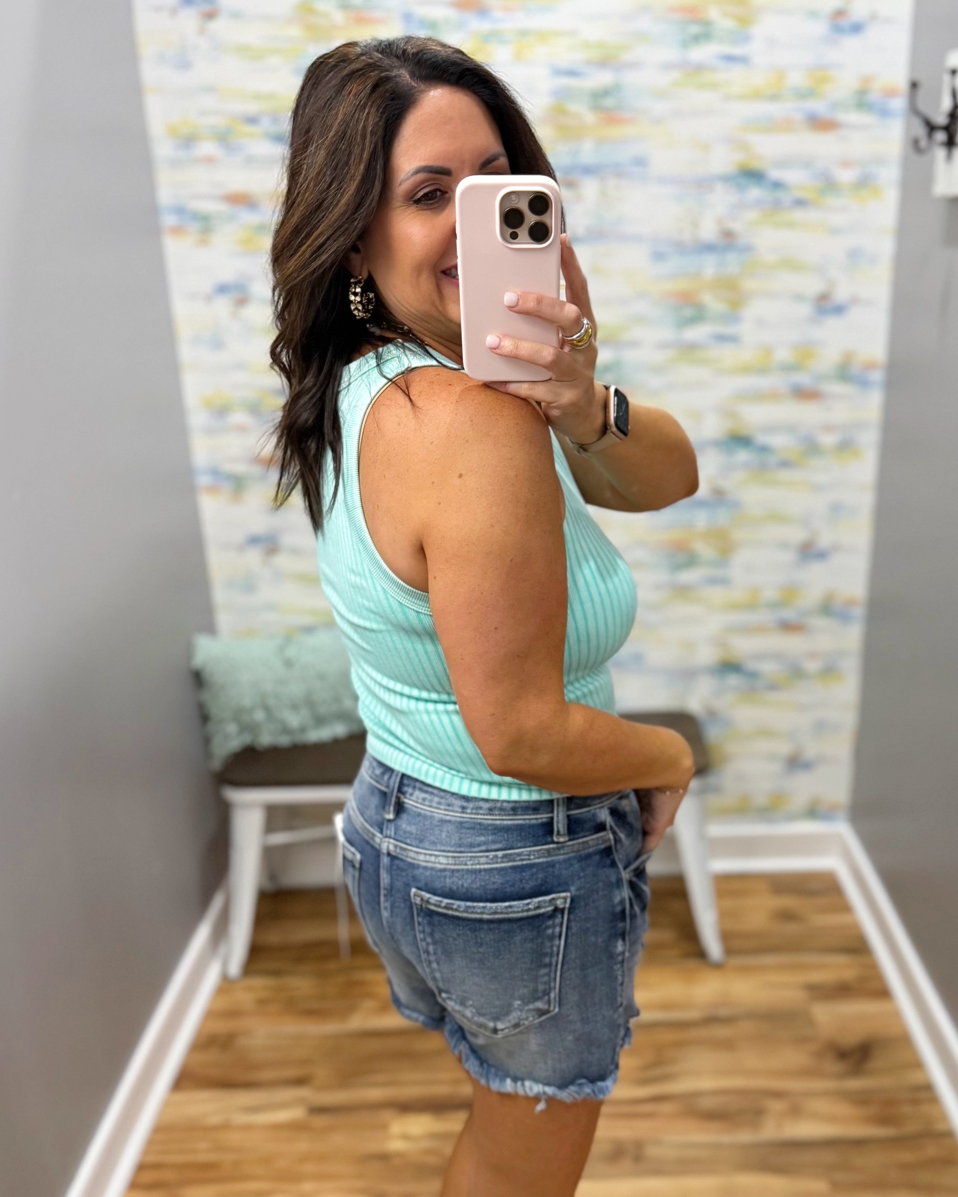 Woman taking a mirror selfie wearing a light blue tank top and denim shorts in a room with a patterned wall.
