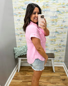 Woman taking a mirror selfie wearing a pink checkered shirt and denim shorts in a room with a textured wall.