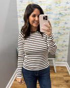 A woman taking a mirror selfie wearing a fitted long-sleeve top with horizontal brown and off-white stripes, paired with dark blue jeans.