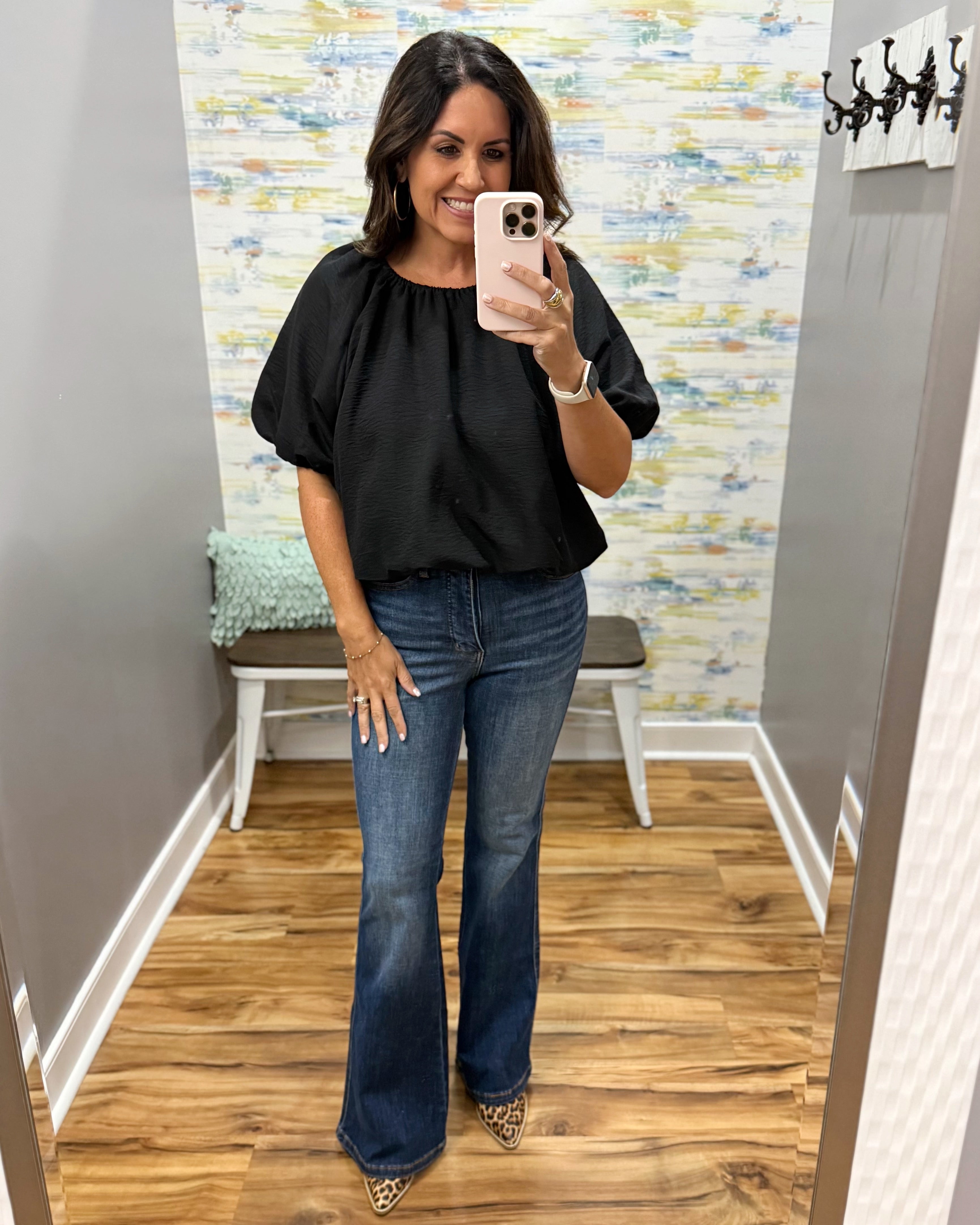 A woman takes a mirror selfie wearing a black puff-sleeve blouse and blue flare jeans in a fitting room with wood floors and a patterned wall.