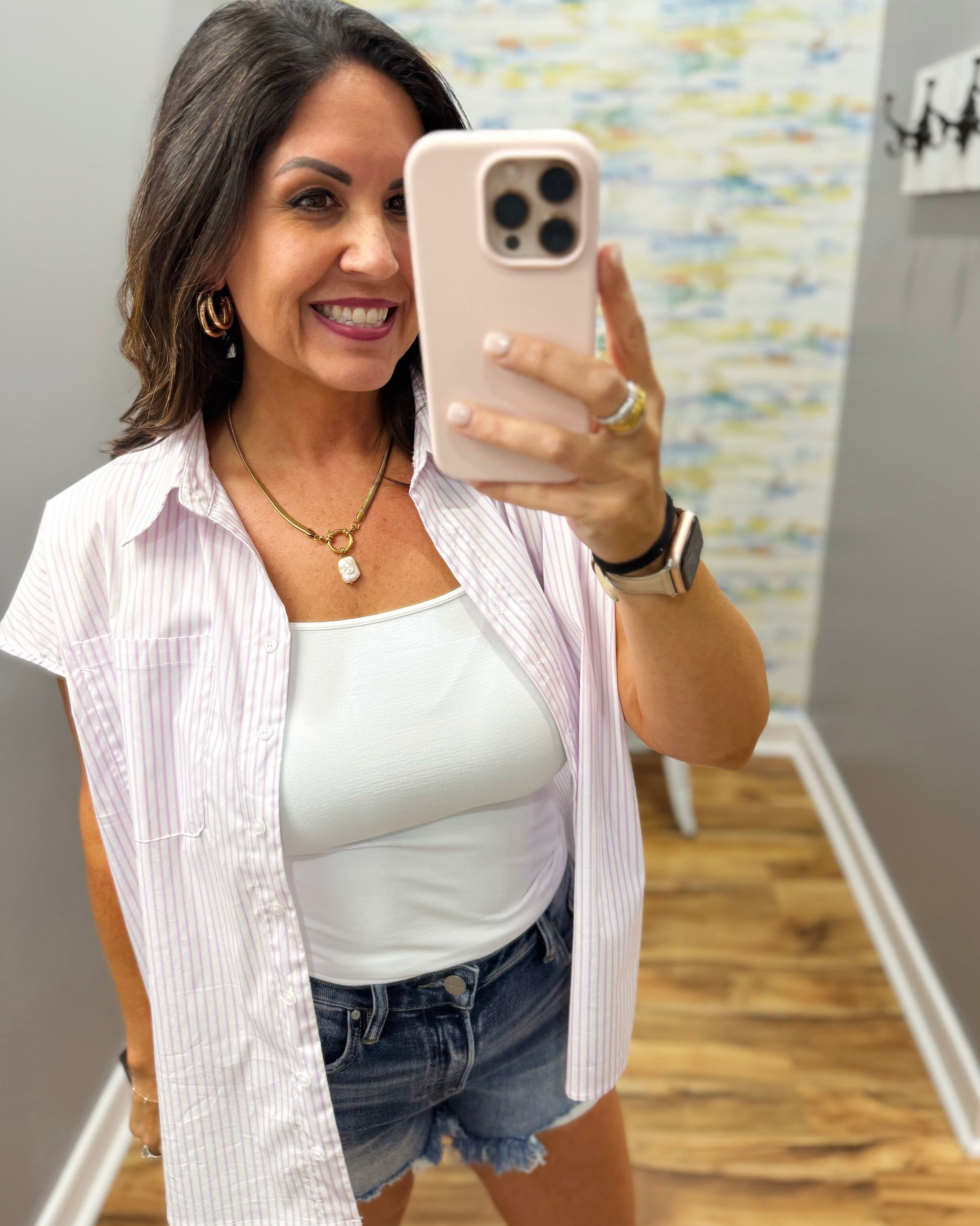 Woman taking a mirror selfie wearing a white shirt over a white tank top and denim shorts.