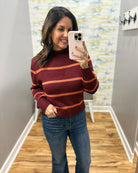 A woman takes a mirror selfie wearing a burgundy and orange striped knit sweater with blue jeans in an indoor hallway.