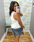Woman taking a mirror selfie wearing a white sleeveless top and denim shorts in a room with colorful wall art.