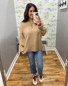 A woman takes a mirror selfie wearing a beige knit crewneck sweater, blue cropped jeans, and white sneakers with black laces in a fitting room with wood floors and a patterned wall.