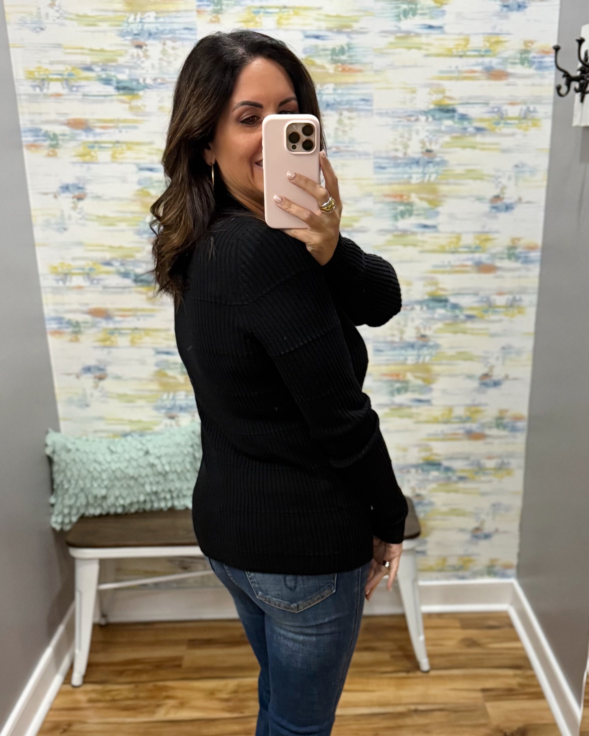 A woman takes a mirror selfie in a fitting room wearing a black ribbed knit long-sleeve sweater and blue jeans, with a patterned wallpaper background and a white bench.