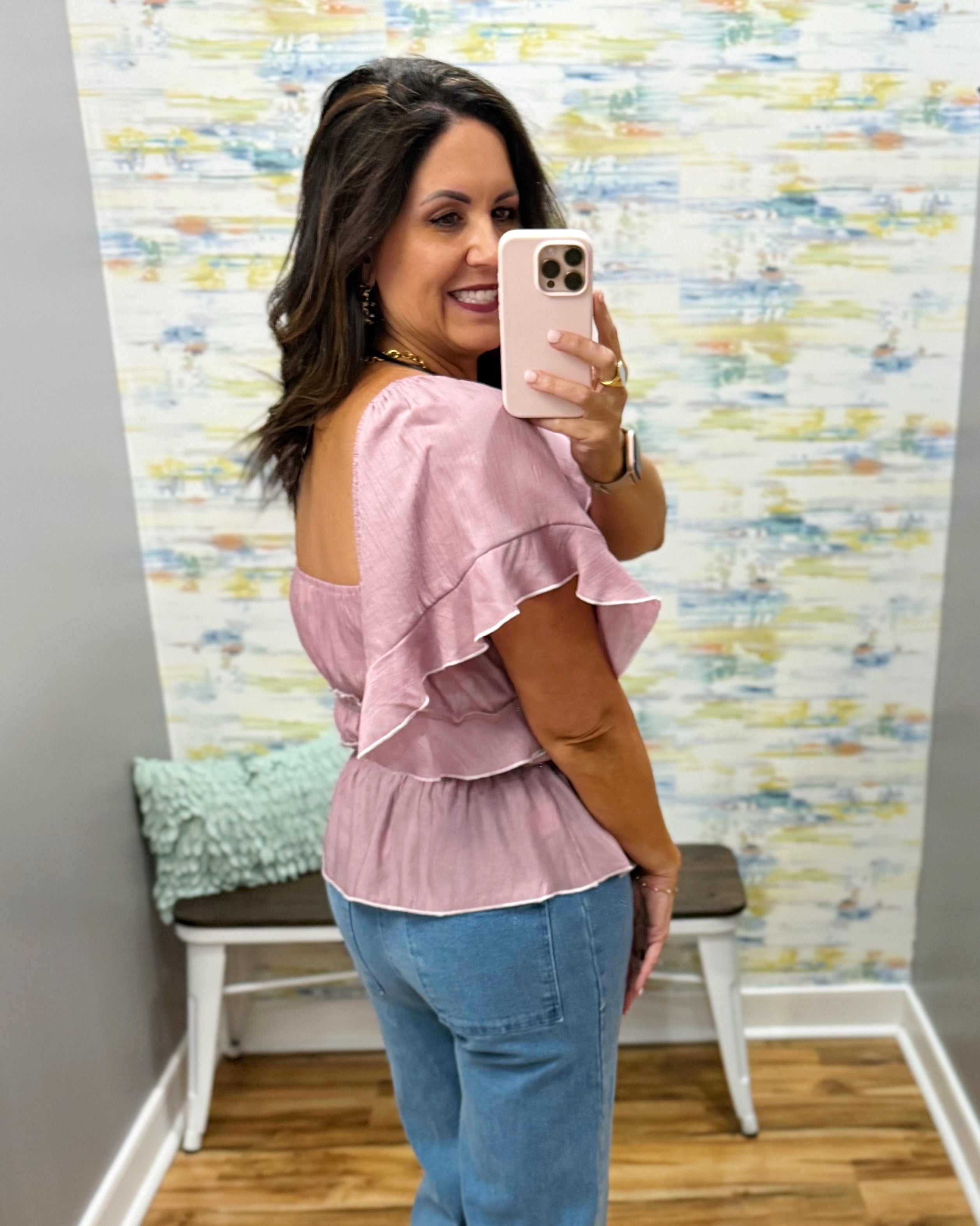 Woman taking a mirror selfie wearing a pink top and blue jeans in front of a colorful wall.