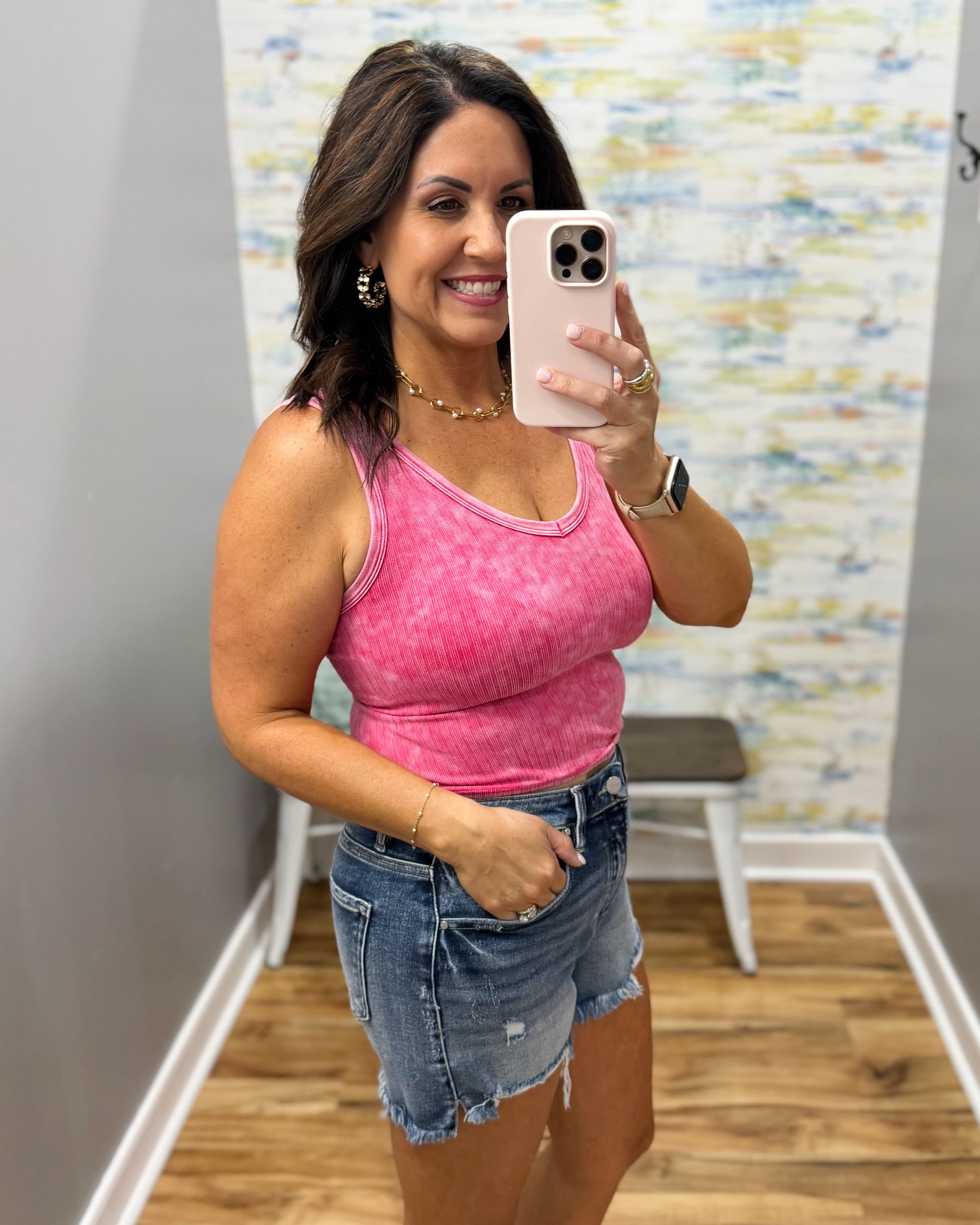 Woman taking a mirror selfie wearing a pink tank top and denim shorts in a room with a colorful wall.