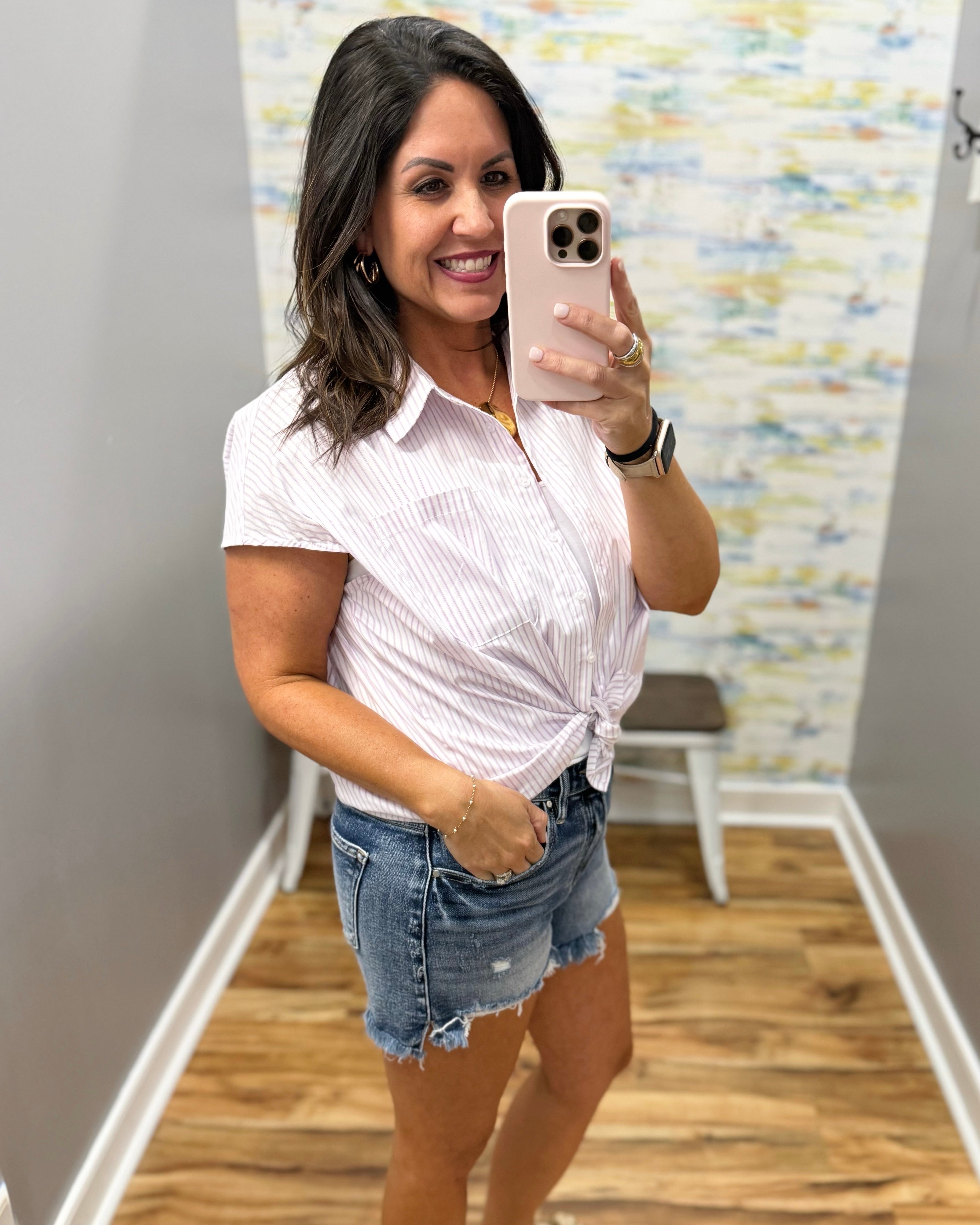 Woman taking a mirror selfie wearing a white short-sleeve shirt and denim shorts in a room with a textured wall.