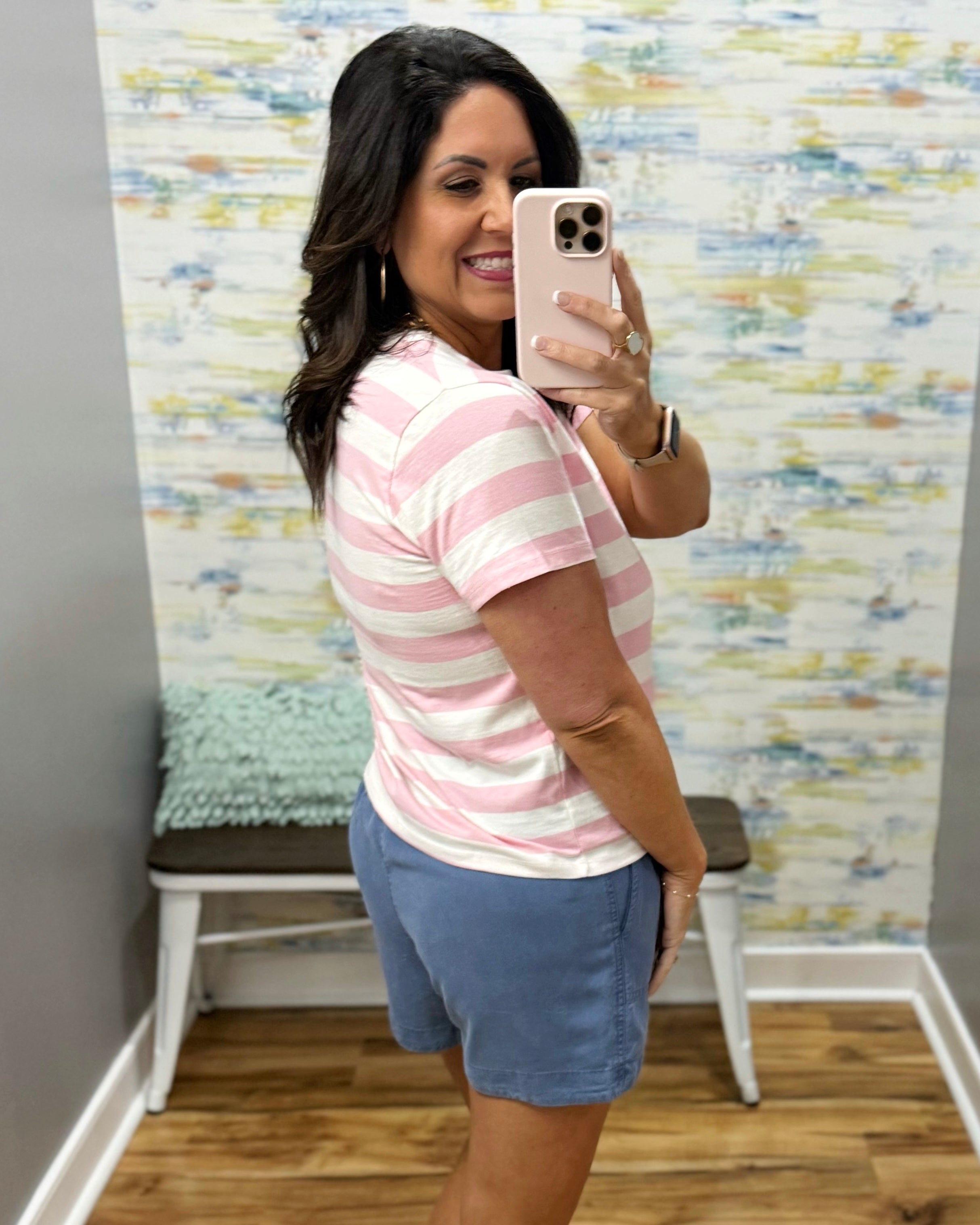 Woman taking a mirror selfie wearing a pink and white striped shirt and blue shorts, standing in a room with a colorful wall.