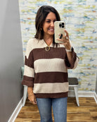 A woman takes a mirror selfie wearing a brown and cream waffle-knit striped sweater with blue jeans, standing in front of a patterned wallpaper background.