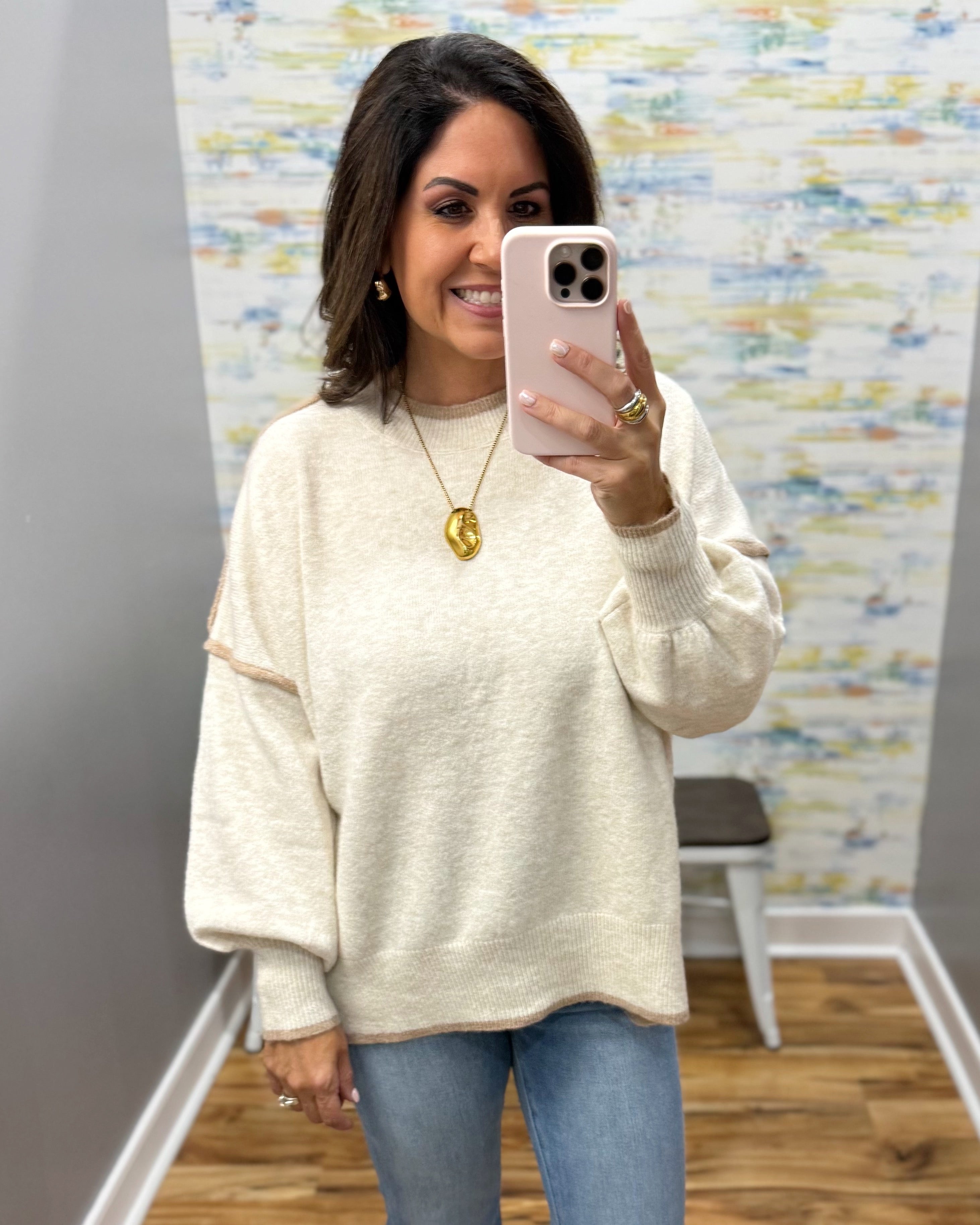 A woman takes a mirror selfie wearing a cream-colored, oversized knit sweater with beige contrast trim on the shoulders and cuffs, paired with blue jeans.