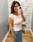 A woman taking a mirror selfie in a fitting room wearing a short-sleeve cowl-neck top with thin peach and white horizontal stripes, paired with blue jeans.