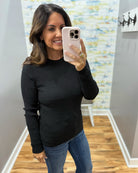 A woman takes a mirror selfie wearing a black ribbed long-sleeve top and blue jeans in an indoor room with wood floors and a patterned wall.