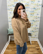 A woman takes a mirror selfie wearing a beige knit crewneck sweater and blue jeans in a room with patterned wallpaper and wood floors.