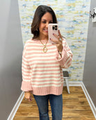 A woman takes a mirror selfie wearing a pink and cream striped knit sweater with blue jeans, standing in a fitting room with a wood floor and abstract wall art.