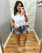 Woman taking a mirror selfie wearing a white shirt and denim shorts in a room with a patterned wall.