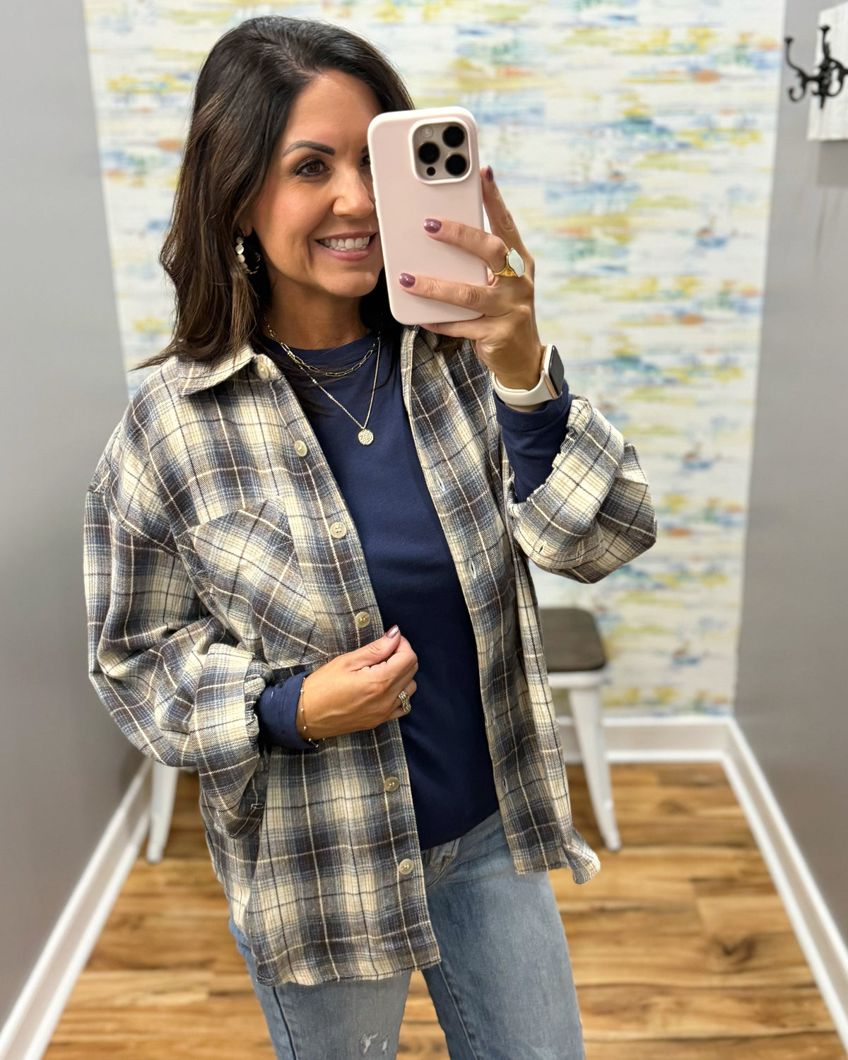 A woman takes a mirror selfie wearing a beige and navy plaid button-up shirt over a navy long-sleeve top, paired with light blue jeans.