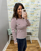 A woman taking a mirror selfie in a fitting room wearing a long-sleeve crewneck top with horizontal burgundy and white stripes, paired with dark blue jeans.