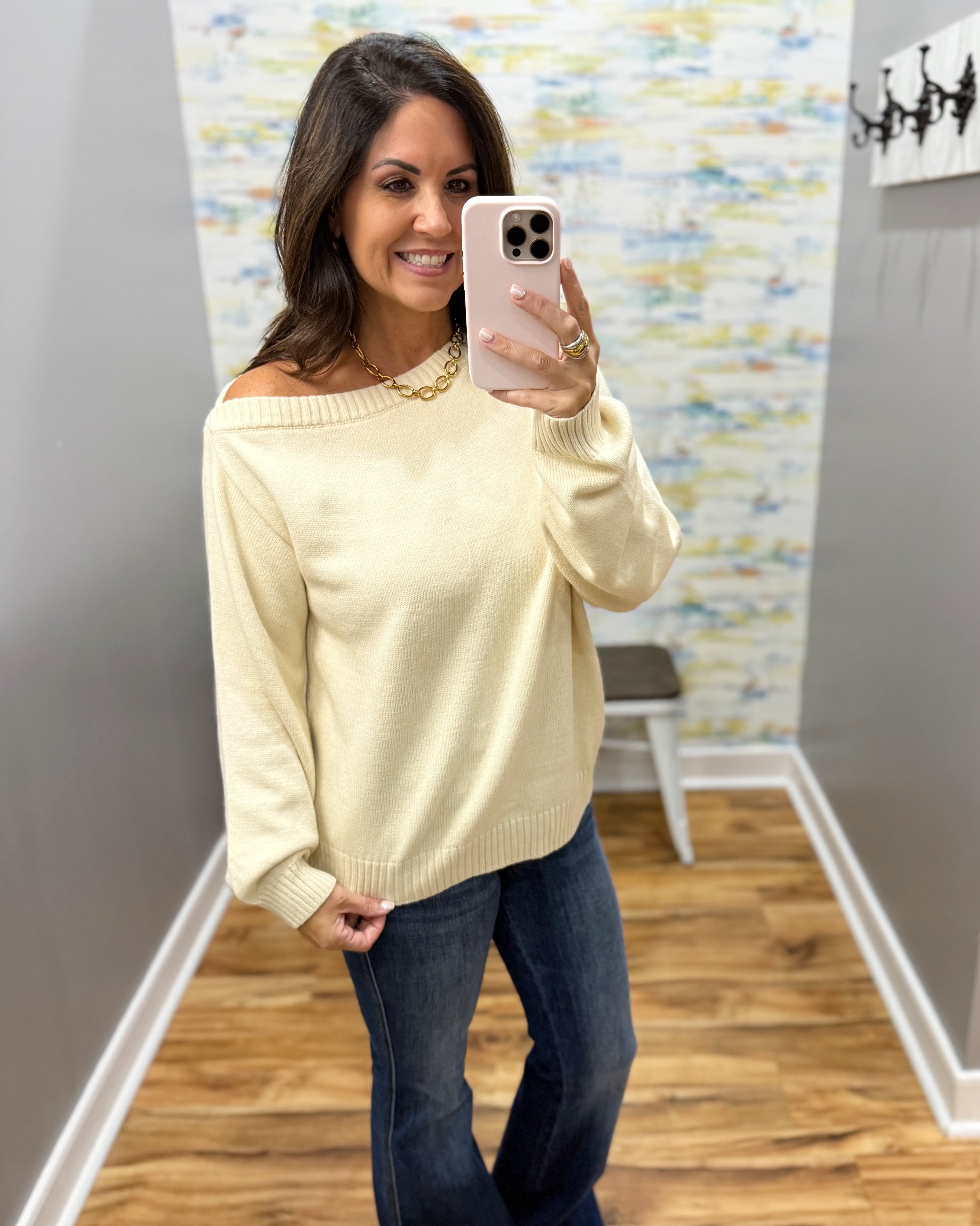 A woman takes a mirror selfie wearing a cream off-the-shoulder knit sweater and dark blue jeans in a home setting with wood floors and a patterned wallpaper background.