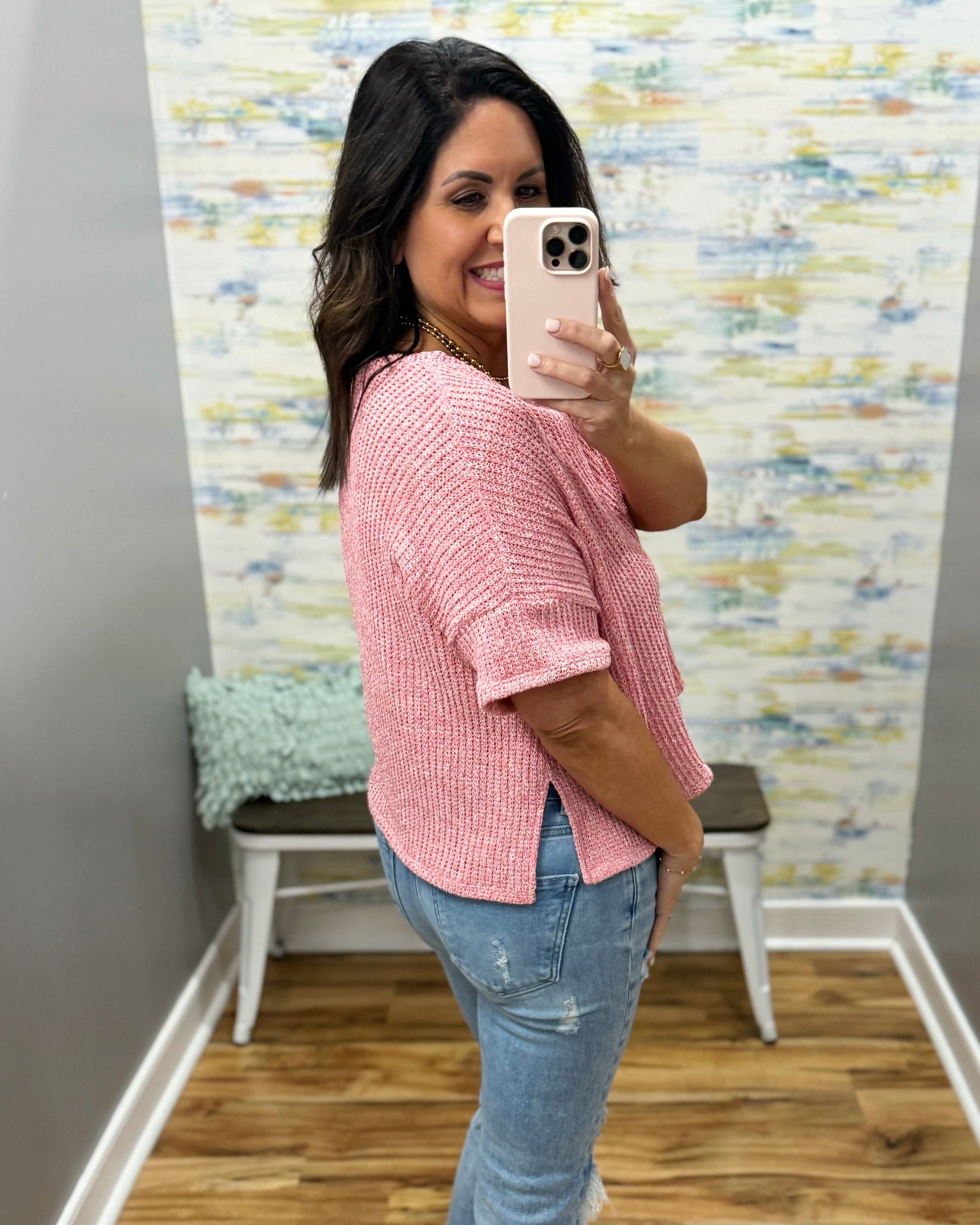 Woman taking a mirror selfie wearing a pink top and ripped jeans in a room with a colorful wall.