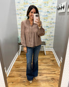 A woman takes a mirror selfie in a fitting room wearing a taupe-brown long-sleeve blouse with pleated detailing and dark blue flare jeans.