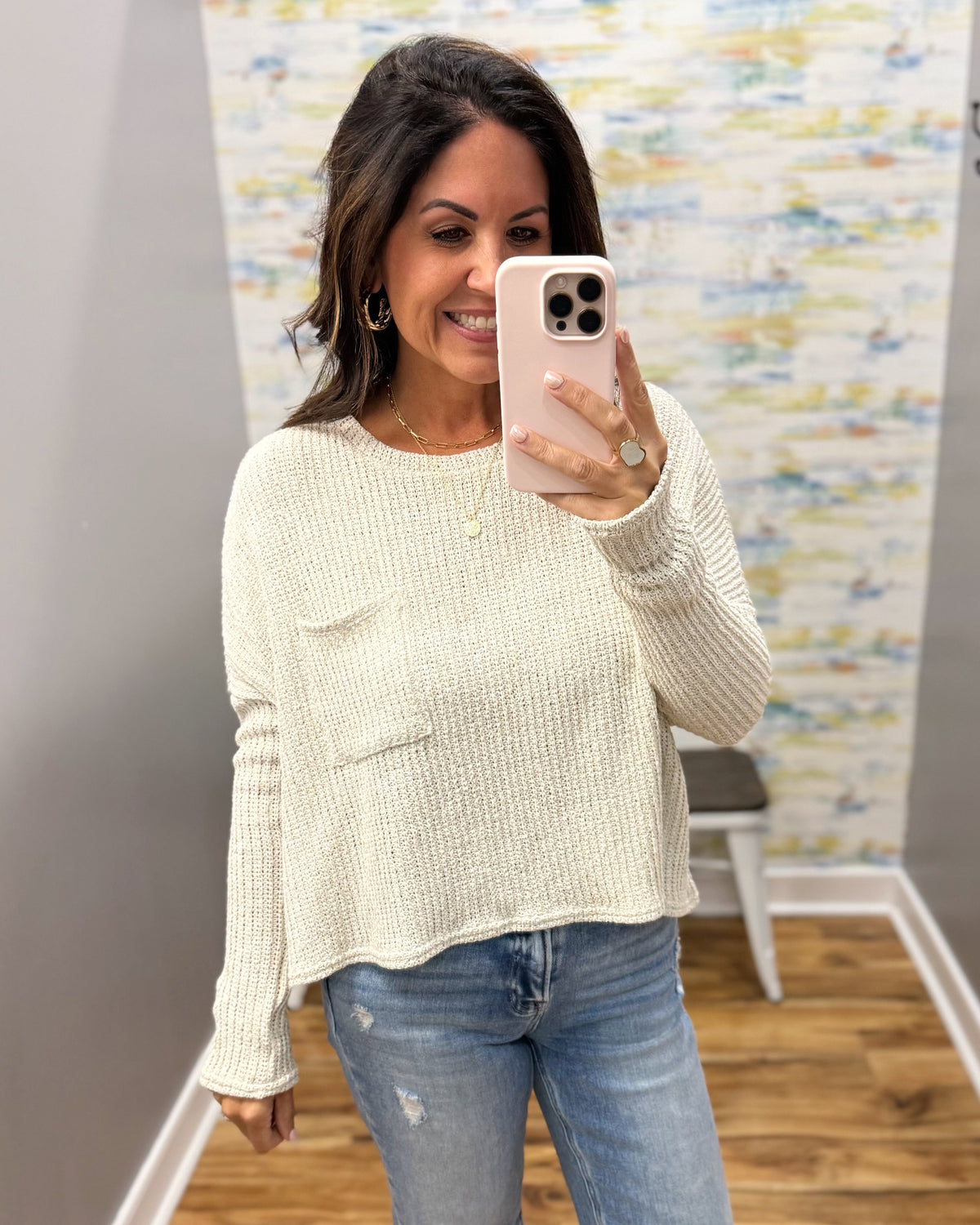 A woman takes a mirror selfie wearing a cream-colored, textured knit long-sleeve sweater with a chest pocket and blue jeans.