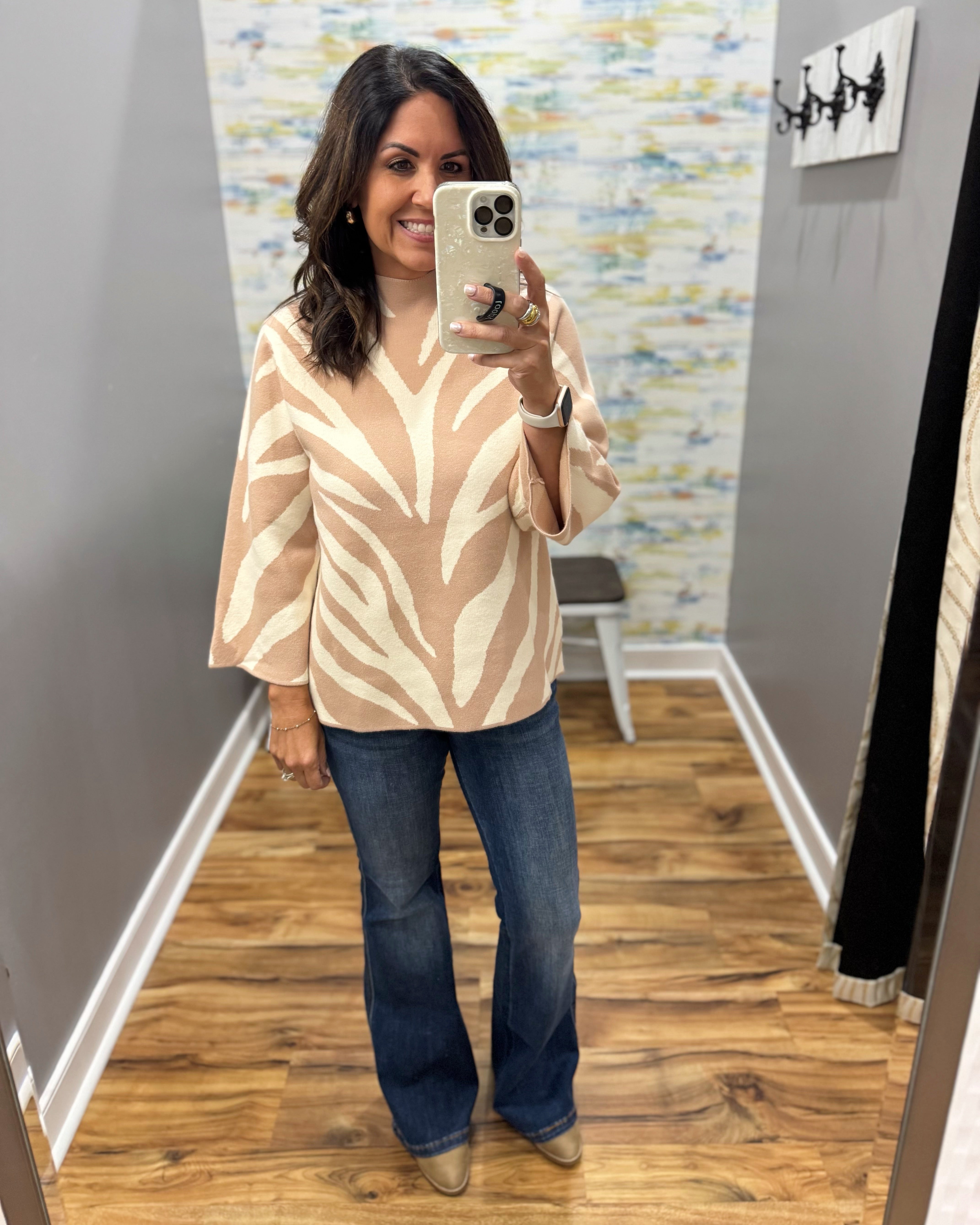 A woman takes a mirror selfie in a fitting room wearing a beige and cream patterned blouse with wide three-quarter sleeves, dark blue flare jeans, and tan pointed-toe flats.