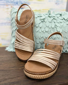 Pair of beige sandals with multiple straps on a wooden surface with a floral-patterned fabric background.