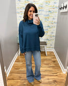A woman takes a mirror selfie in a fitting room wearing a dark teal blue turtleneck sweatshirt and light-wash blue flare jeans with tan pointed-toe flats.