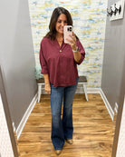 A woman takes a mirror selfie in a fitting room wearing a burgundy button-down shirt with rolled sleeves and blue flare jeans.