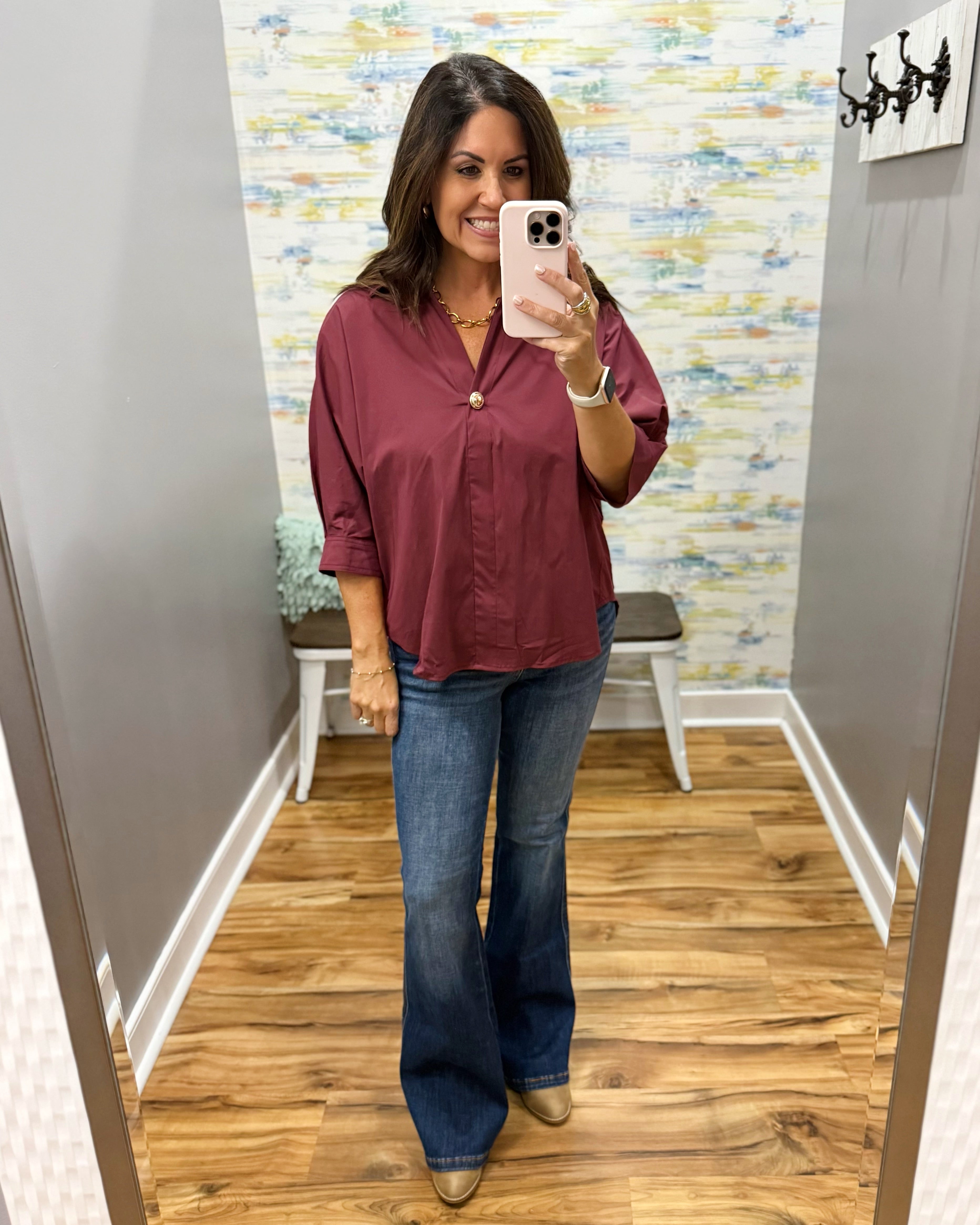 A woman takes a mirror selfie in a fitting room wearing a burgundy button-down shirt with rolled sleeves and blue flare jeans.