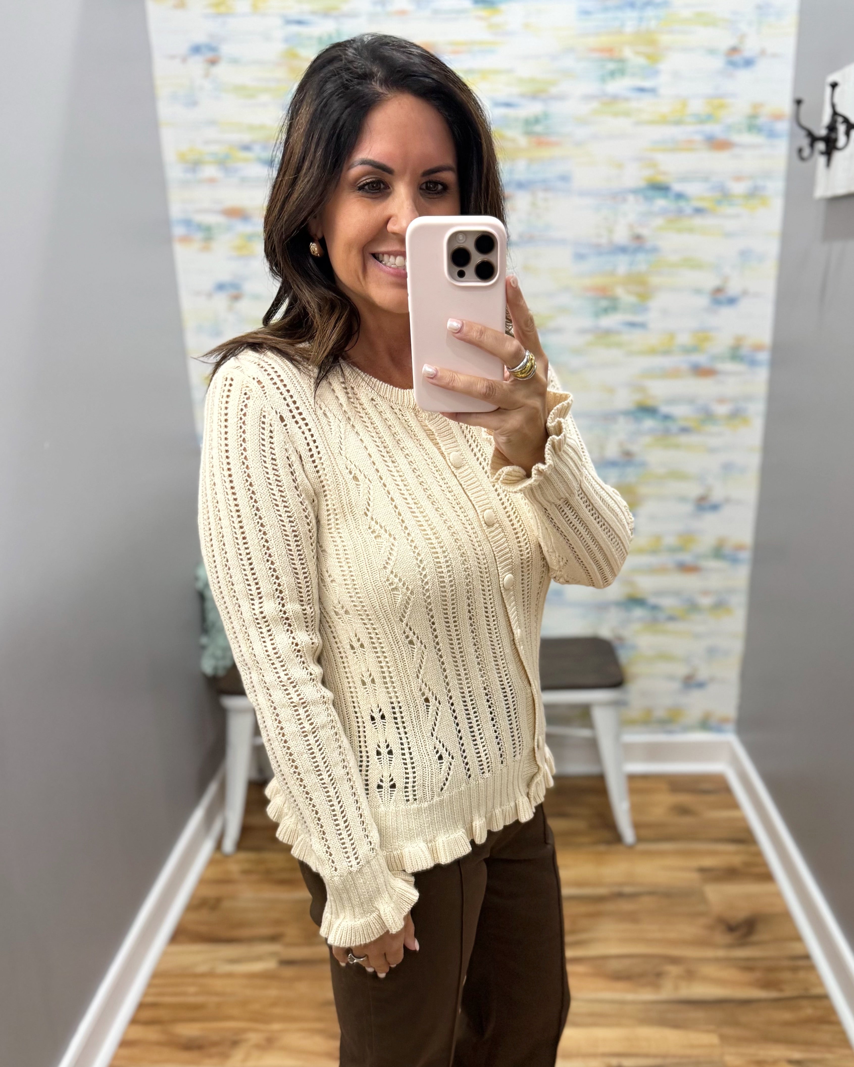 A woman takes a mirror selfie wearing a cream-colored, long-sleeve knit cardigan sweater with a ruffled hem and cuffs, paired with brown pants in a home hallway setting.