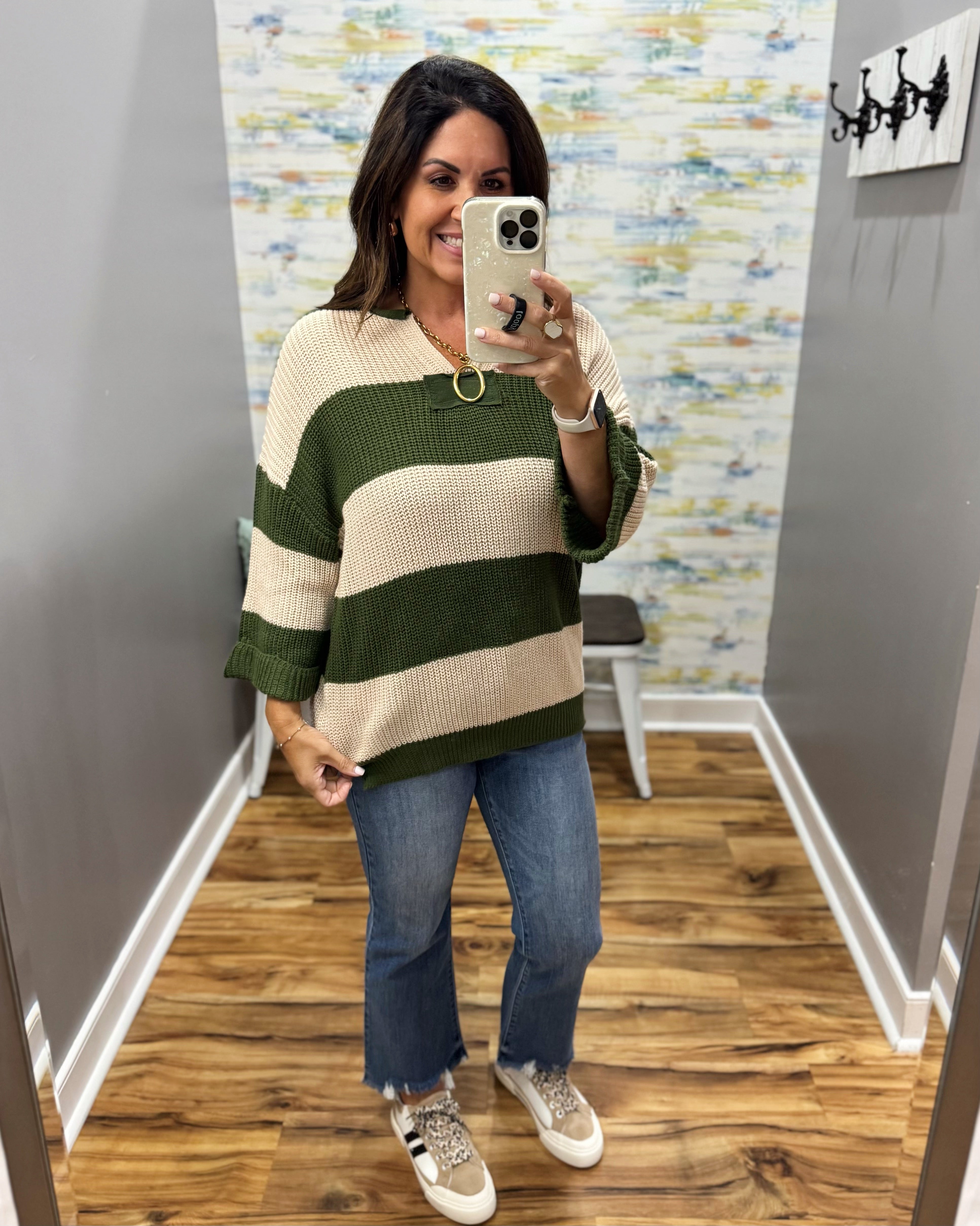A woman takes a mirror selfie wearing a green and cream striped knit sweater, blue jeans, and white sneakers in a fitting room with wood floors and a patterned wall.