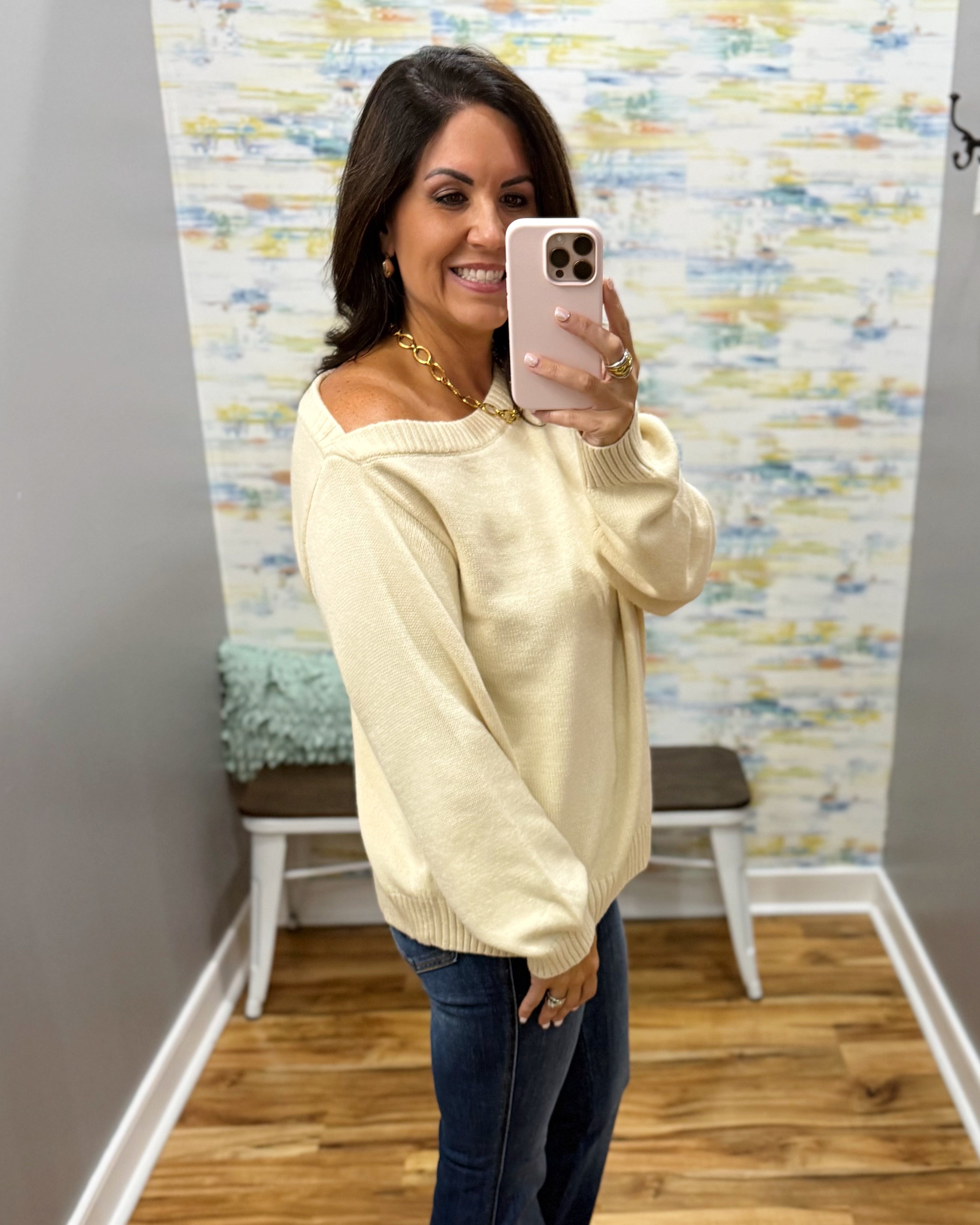 A woman takes a mirror selfie wearing a cream-colored off-the-shoulder knit sweater and dark blue jeans in a fitting room with a patterned wallpaper background.