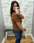 A woman takes a mirror selfie in a fitting room wearing a brown short-sleeve knit top with textured side panels and blue jeans.