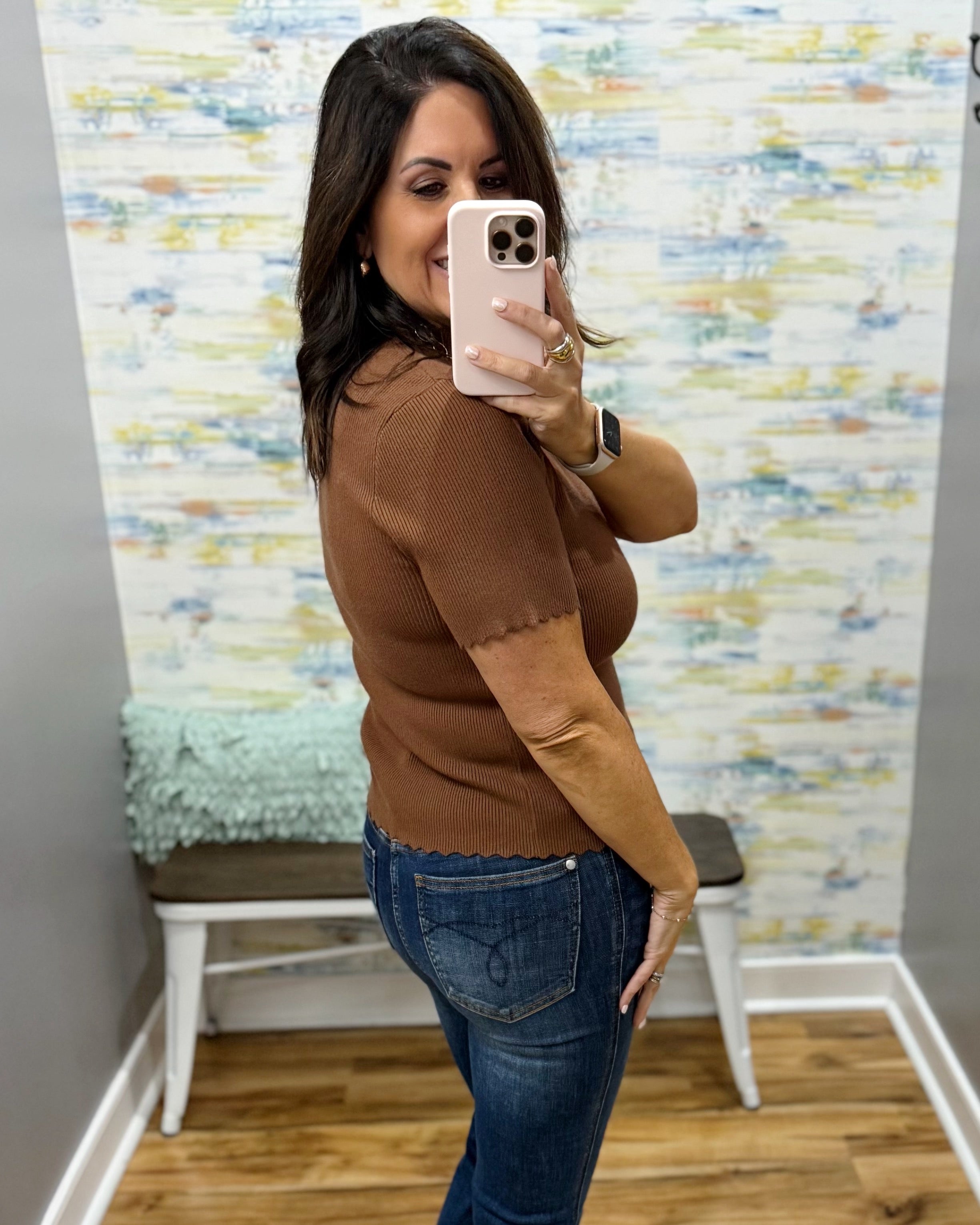 A woman takes a mirror selfie in a fitting room wearing a brown short-sleeve knit top with textured side panels and blue jeans.