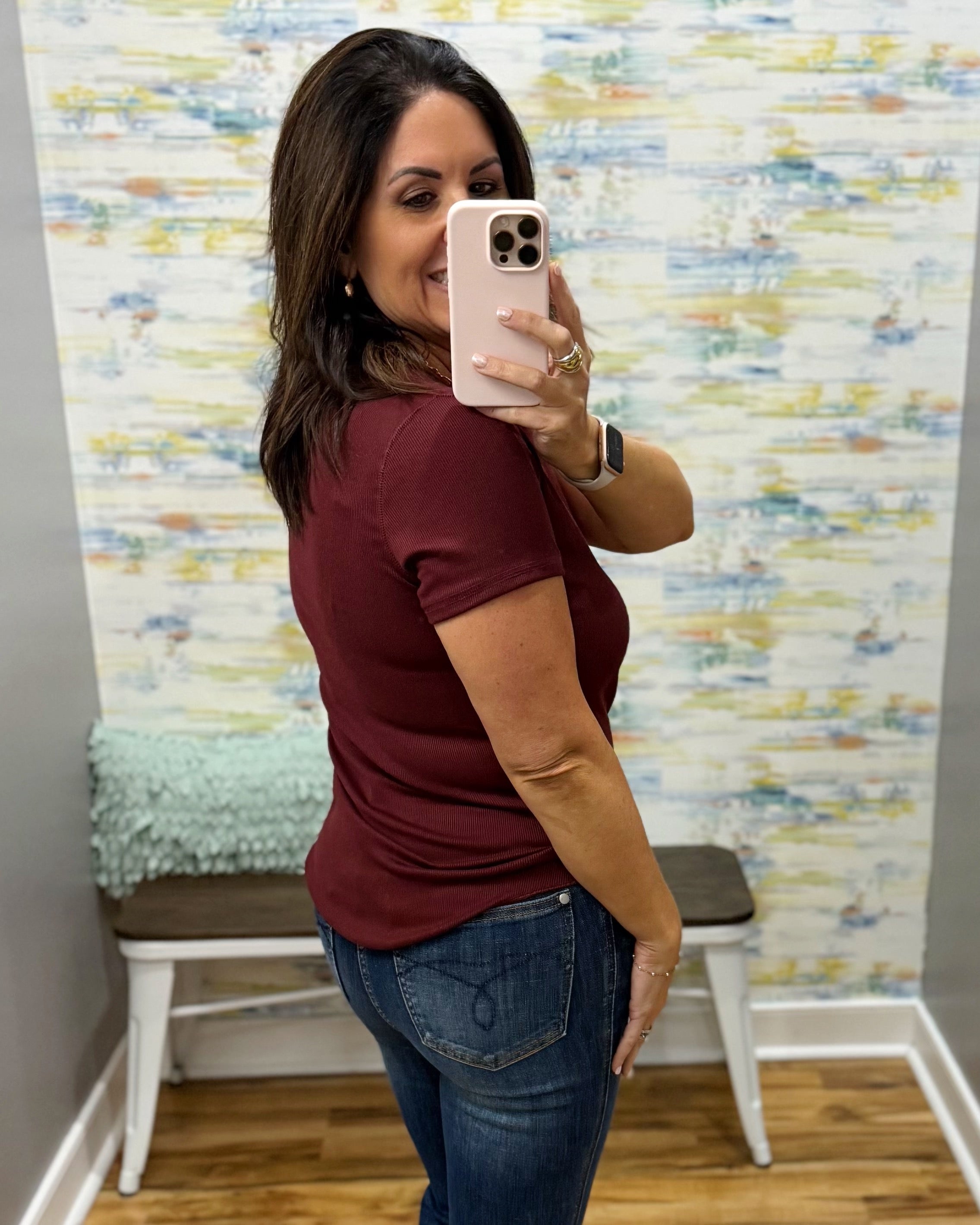 A woman taking a mirror selfie wearing a short-sleeve maroon t-shirt and blue jeans, standing in front of a patterned wallpaper background with a bench.