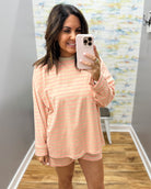 A woman takes a mirror selfie in a fitting room wearing a peach and white striped long-sleeve t-shirt and matching shorts, paired with beige slide sandals.