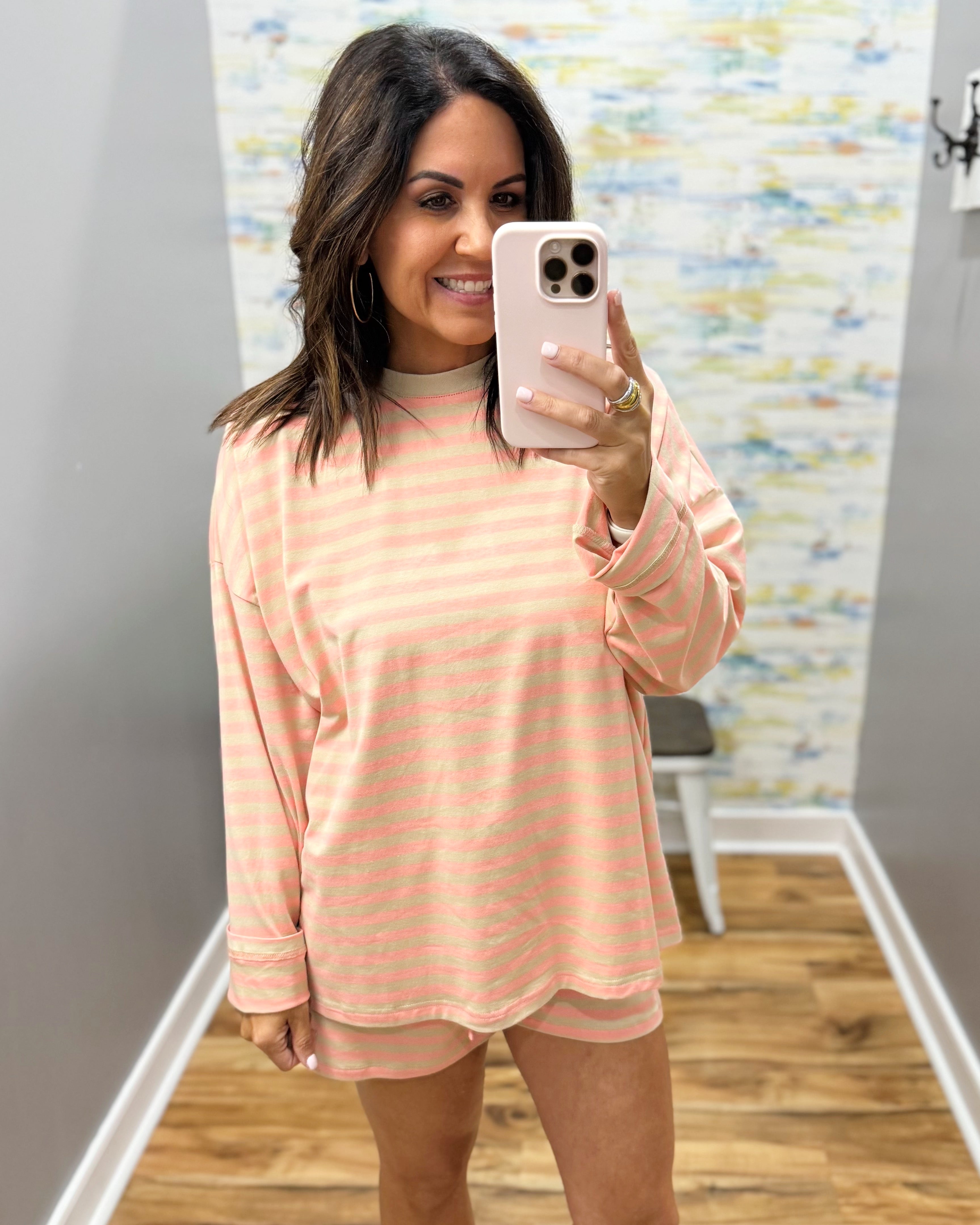 A woman takes a mirror selfie in a fitting room wearing a peach and white striped long-sleeve t-shirt and matching shorts, paired with beige slide sandals.
