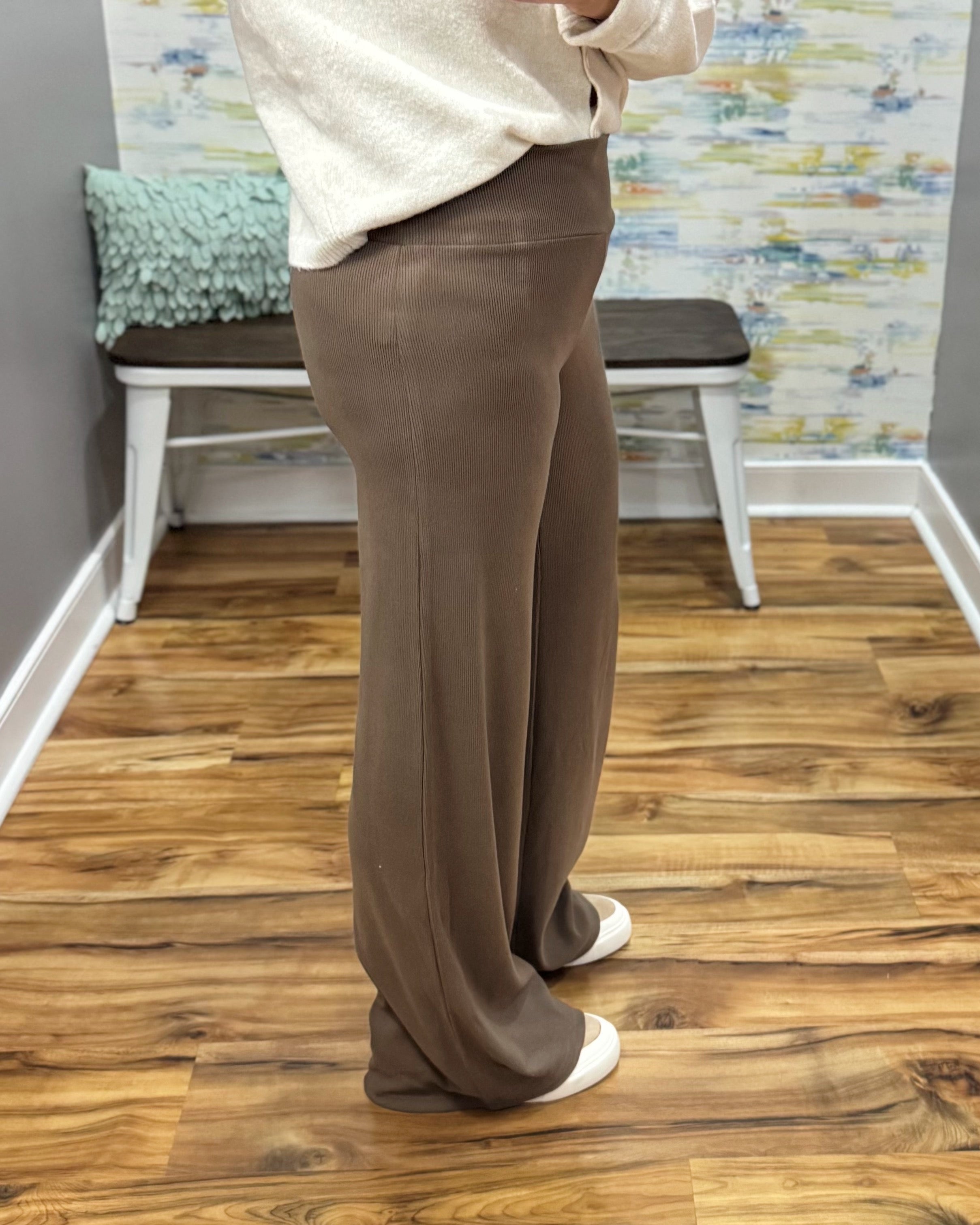 A person wearing high-waisted, wide-leg brown pants, a cream-colored top, and white slip-on shoes, standing on a wood floor in front of a bench and patterned wall.