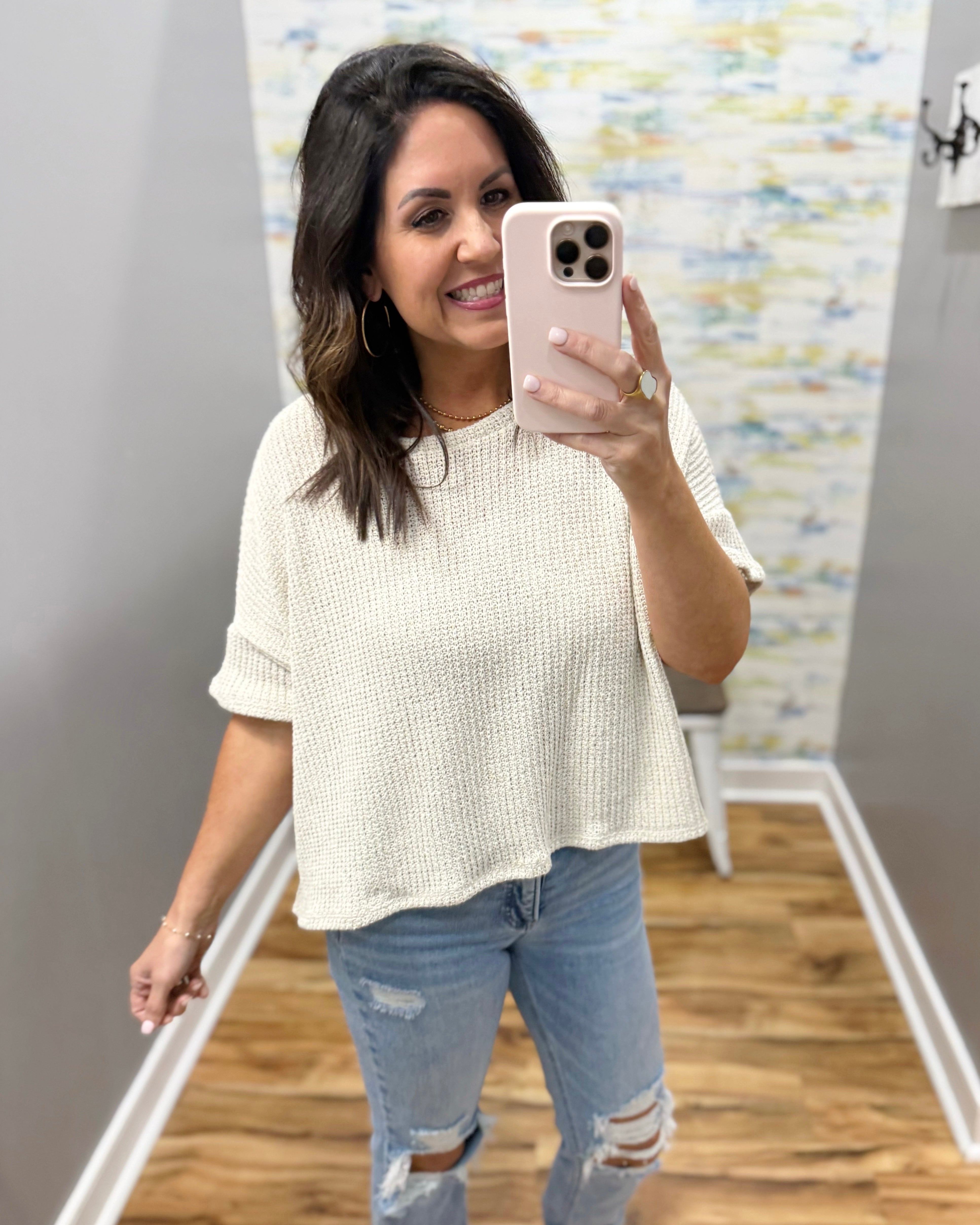 Woman taking a mirror selfie wearing a natural sweater and jeans in a room with a textured wall.