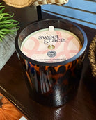 A Sweet Grace scented soy candle in a dark tortoiseshell-patterned glass jar, sitting on a dark wooden table with a green plant and woven basket in the background.
