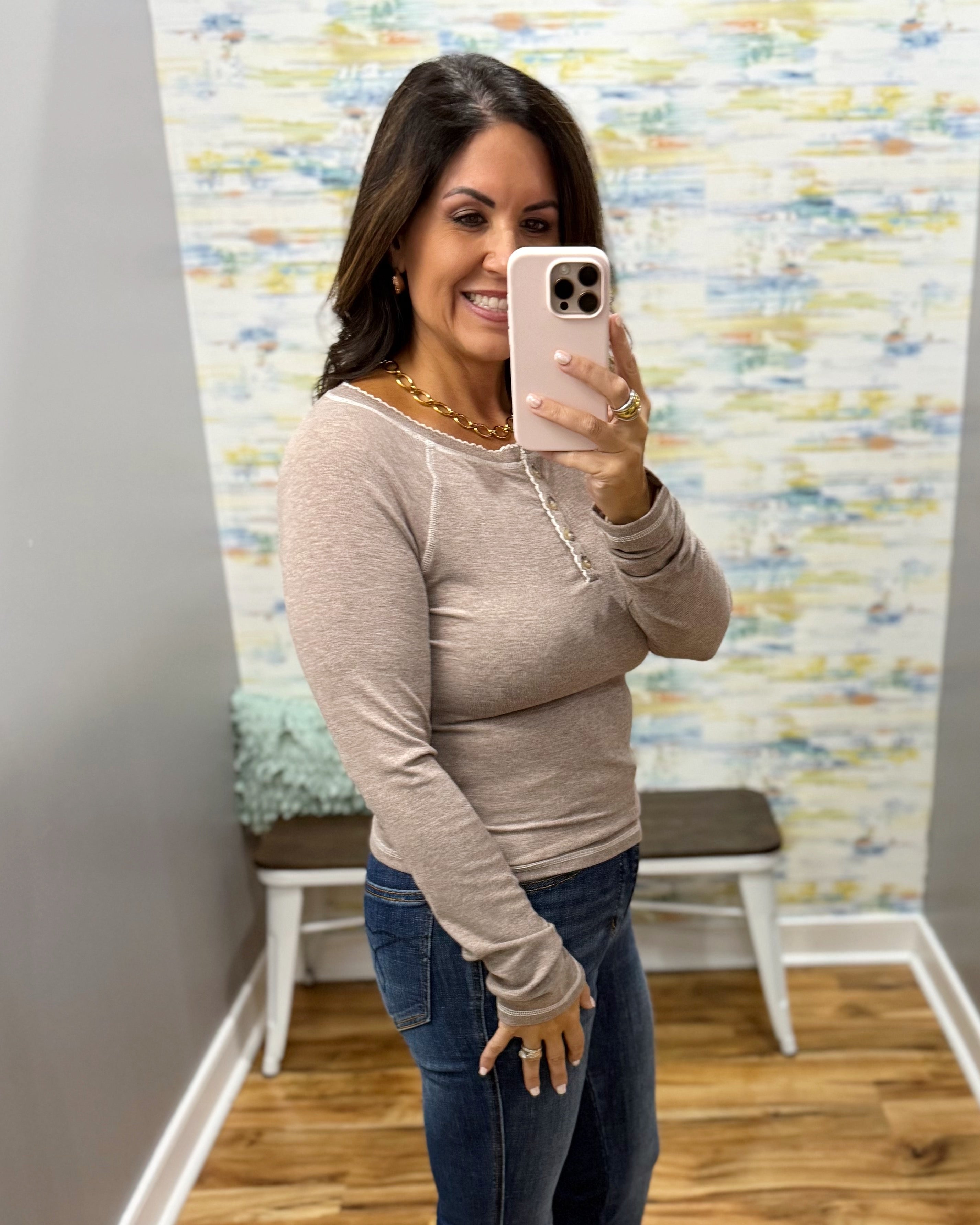 A woman takes a mirror selfie wearing a beige long-sleeve top with lace-up front details and blue jeans in a fitting room with a patterned wallpaper background.